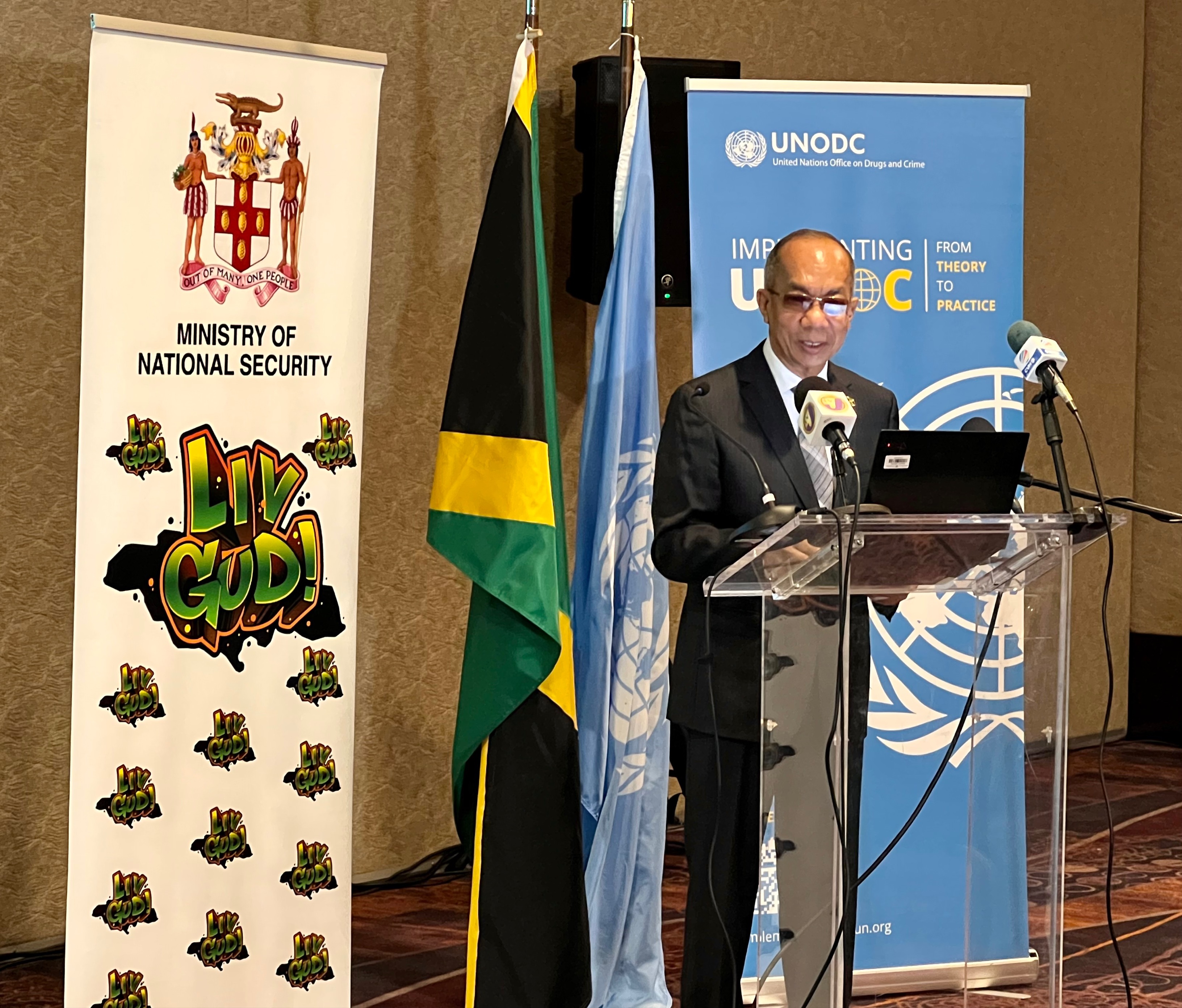 A man speaking behind a podium. On the left side, the roll up banner of Jamaica's Ministry of National Security. Behind him, the flags of Jamaica and of the United Nations, and the roll up banner of the Global Programme on Implementing the Organized Crime Convention: from Theory to Practice. 