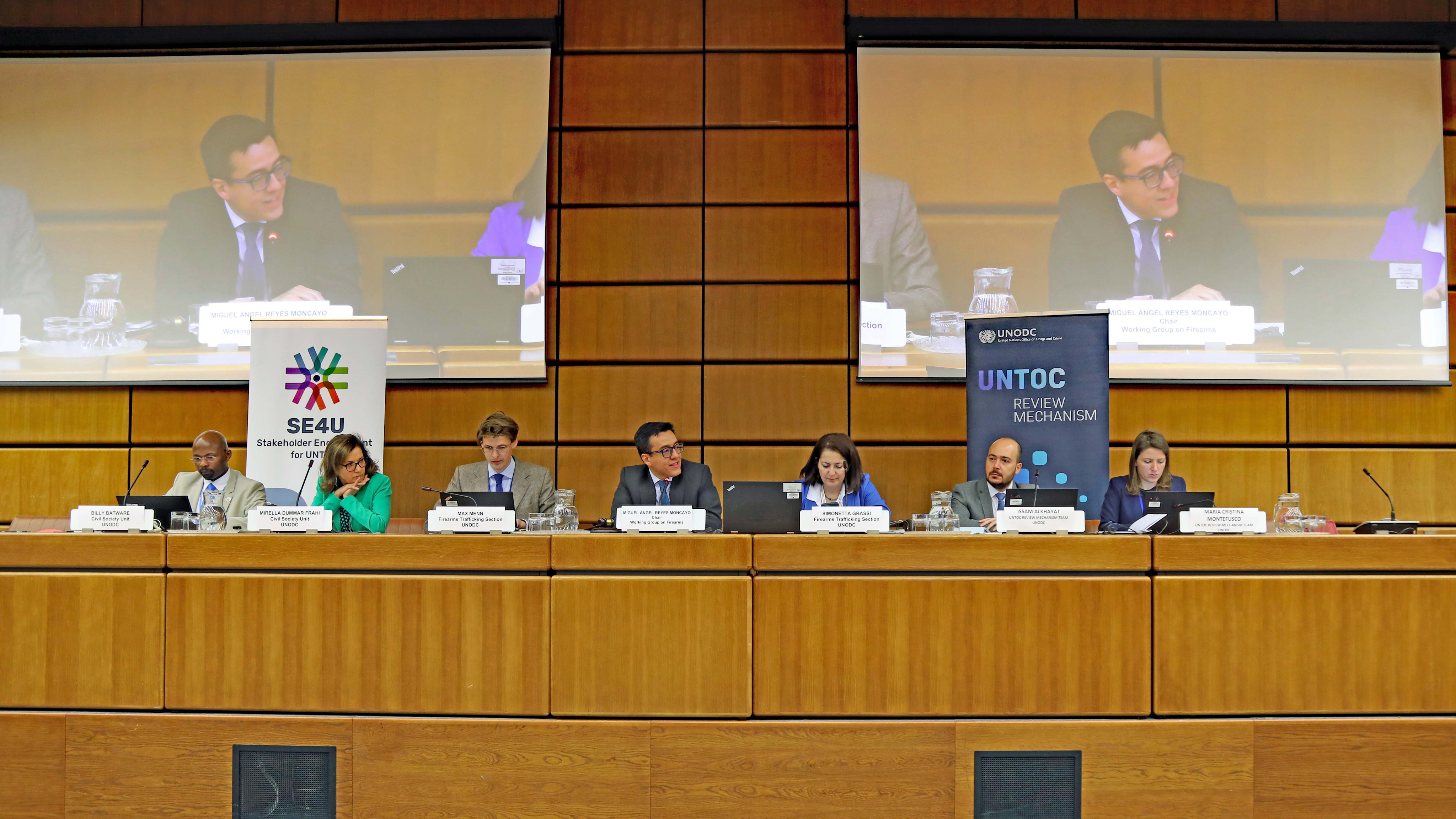 Panel with three women and four men, and their name plates. On the wall, two projector screens display the image of a man speaking who is sitting at the center of the panel. Behind the panel, on the left side, a roll up banner with the logo of SE4U Stakeholder Engagement for UNTOC and on the right side, a roll up banner with the logo of the UNTOC Review Mechanism.