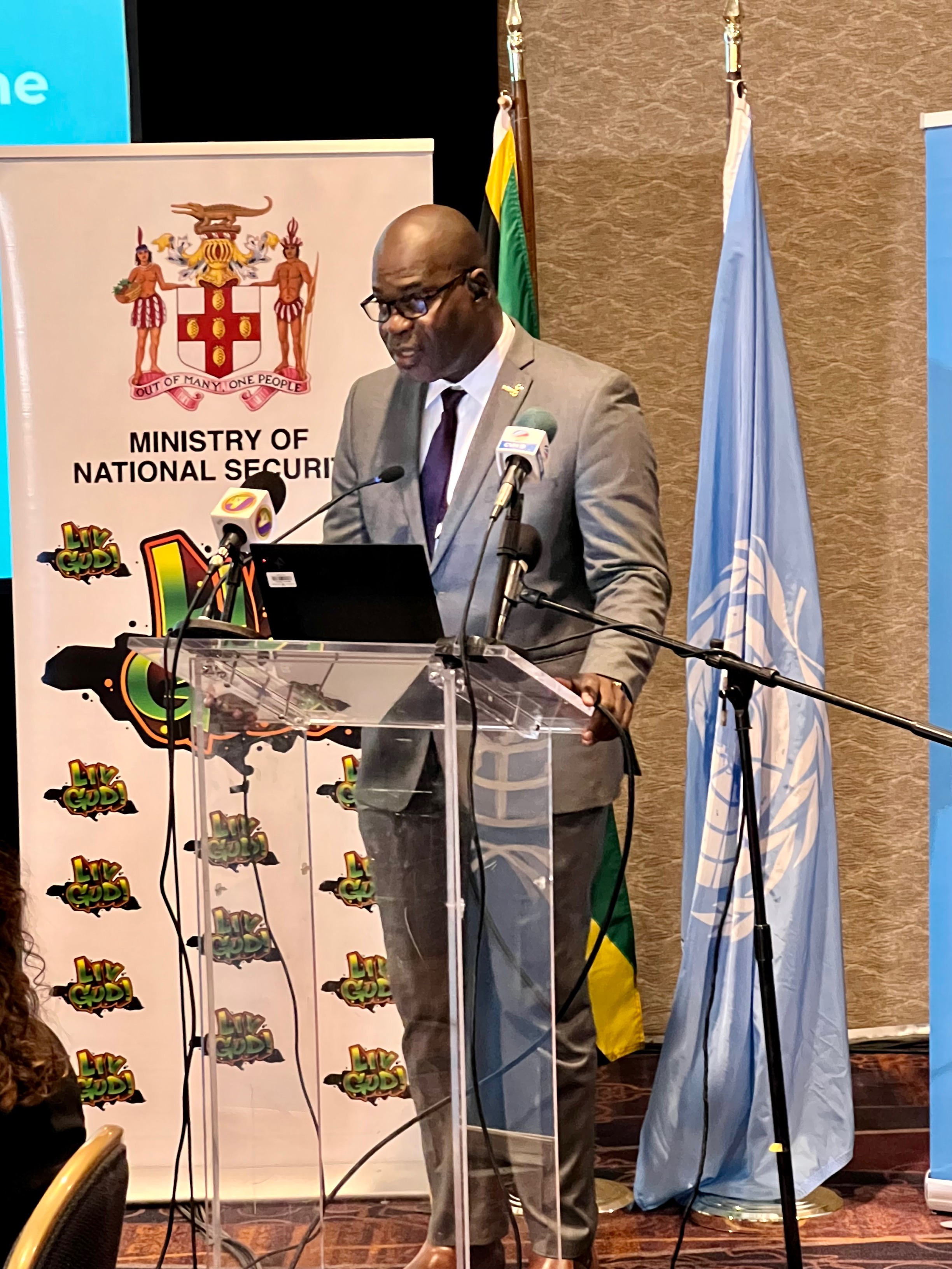 A man speaking behind a podium. On the left side, the roll up banner of Jamaica's Ministry of National Security. Behind him, the flags of Jamaica and of the United Nations.