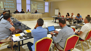 Participants of a meeting are sitting behind a U-shaped table. In the background, a panel with three men and a woman. Behind the panel, two projector screens display the logo of Hotel Mepas.