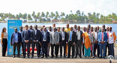 A group photo with participants of a meeting taken outside. On the left side of the photo, the roll up banner of the Global Programme on Implementing the Organized Crime Convention is visible. In the background, palm trees, several houses and the sea.