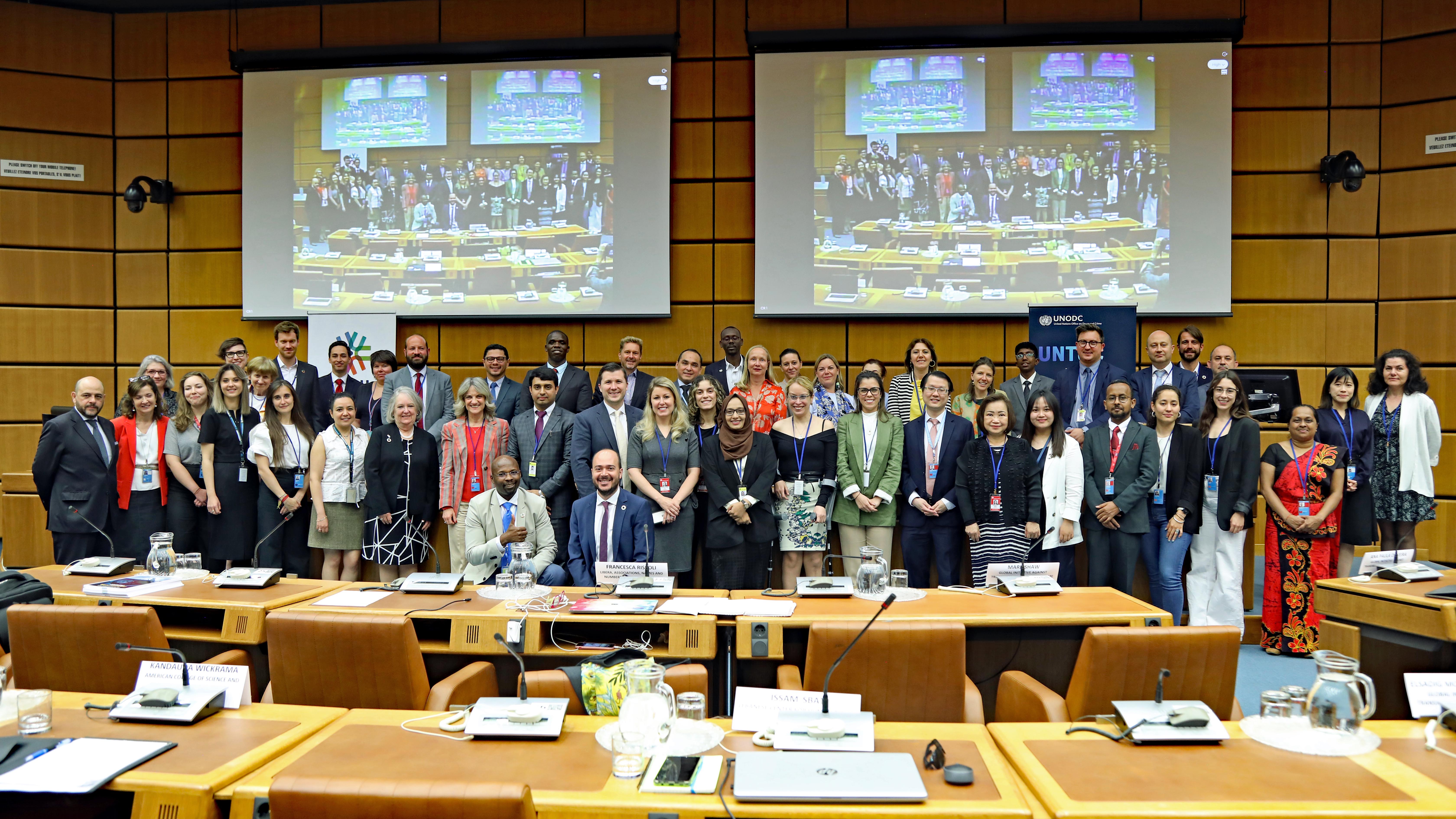 A group photo taken in a meeting room with participants of a meeting. On the wall, two projector screens display an image of the room. Behind the participants, two roll up banners: one of the UNTOC Review Mechanism and the other of Stakeholder Engagement for UNTOC (SE4U).