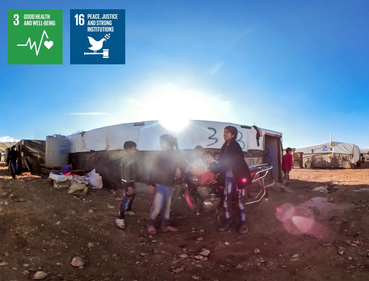 Children playing outside of a refugee tent with the logos of SDG 3 and SDG 16 featured on the side. 