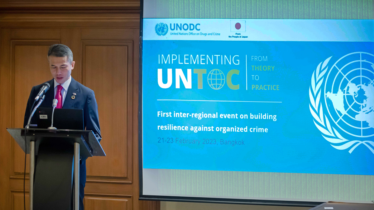 A man is speaking behind a podium. On the wall, a projector screen with the logo of UNODC and of the Global Programme on Implementing the Organized Crime Convention: from Theory to Practice, and the flag of Japan with the words "From the People of Japan". The following text appears on the projector screen: "First inter-regional event on building resilience against organized crime - 21-23 February 2023, Bangkok".