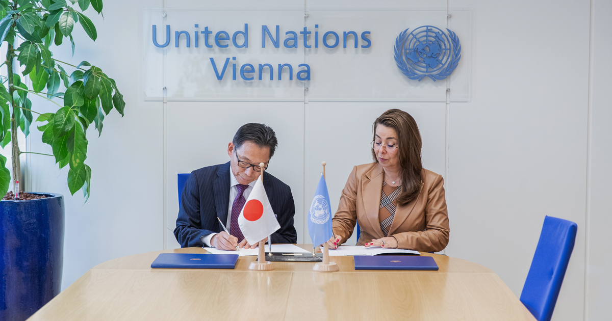 The Ambassador of Japan and UNODC’s Executive Director signing the agreement at a table adorned with the flags of Japan and the United Nations.  