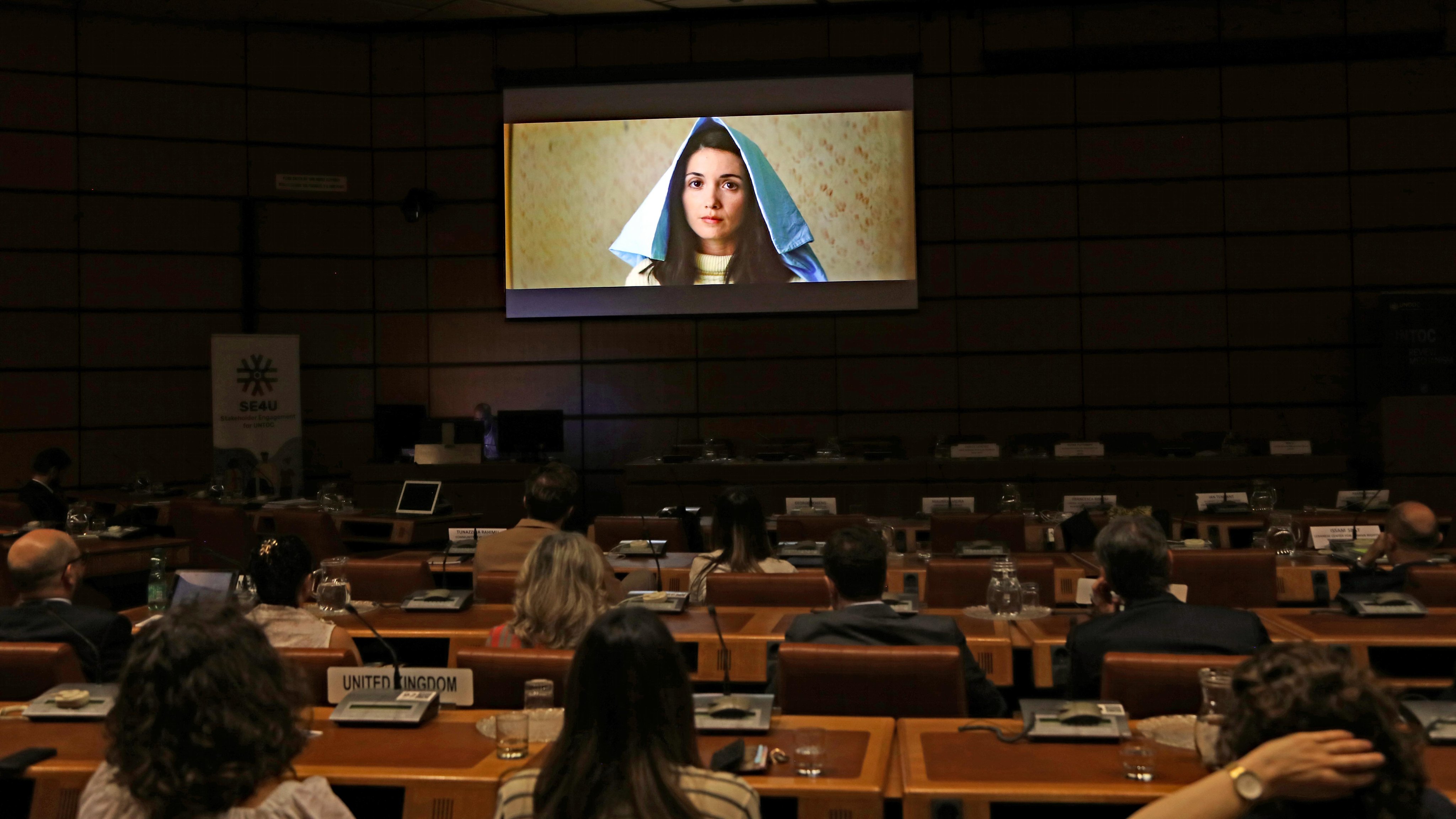 A projector screen displays an image from a film. Participants of a film screening are sitting behind tables.
