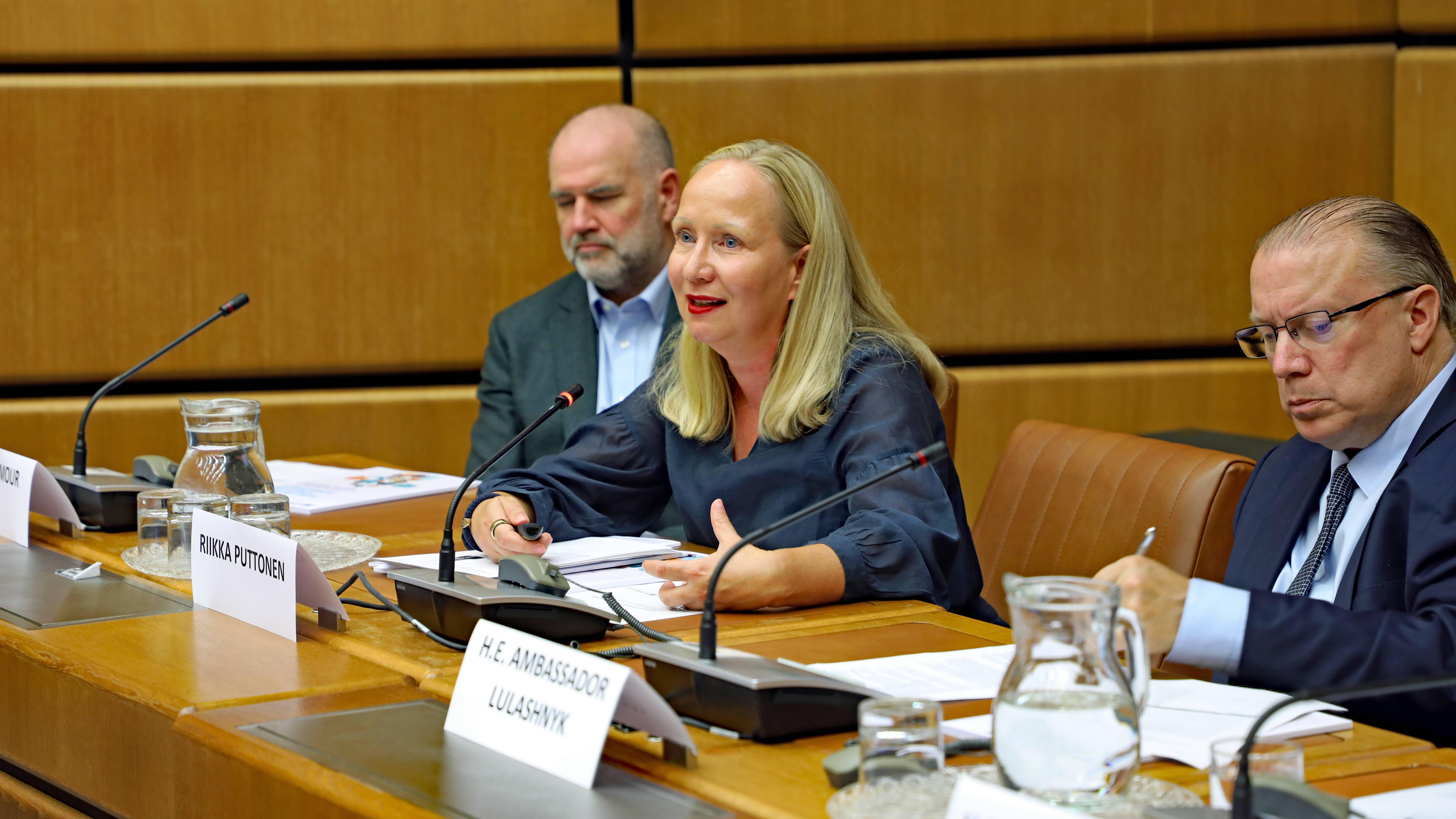 A panel with two men and a woman. Their name plates are visible on the table. One of the panellists, a woman, is speaking through a microphone.