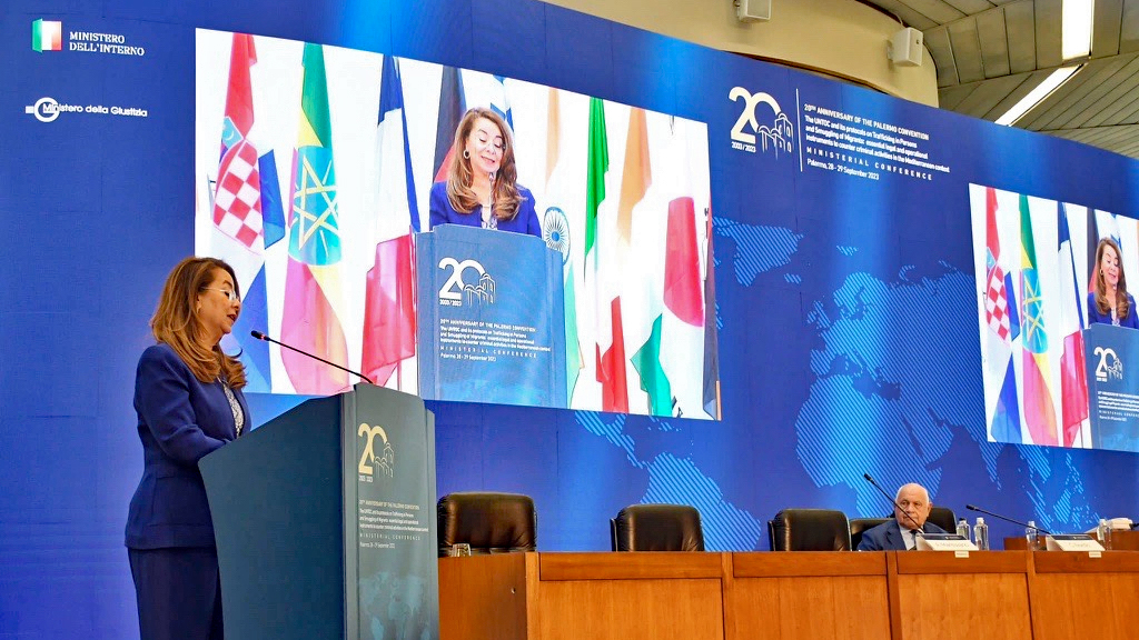A woman is speaking behind a lectern through a microphone. In the background, a screen displays two images of the woman speaking. On the right side of the photo, a man is sitting behind a table.