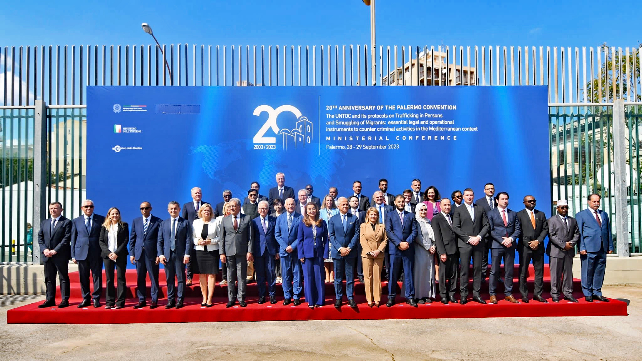 A group photo taken outside a building arranged in three rows. In the background, a banner with text reading "20th Anniversary of the Palermo Convention - The UNTOC and its protocols on Trafficking in Persons and Smuggling of Migrants: essential legal and operational instruments to counter criminal activities in the Mediterranean context - Ministerial Conference - Palermo, 28-29 September 2023".