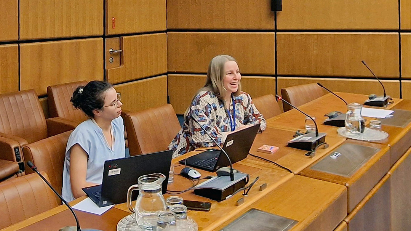 A panel with two women. The woman on the right side is speaking on a microphone. Two laptops are visible on the table.