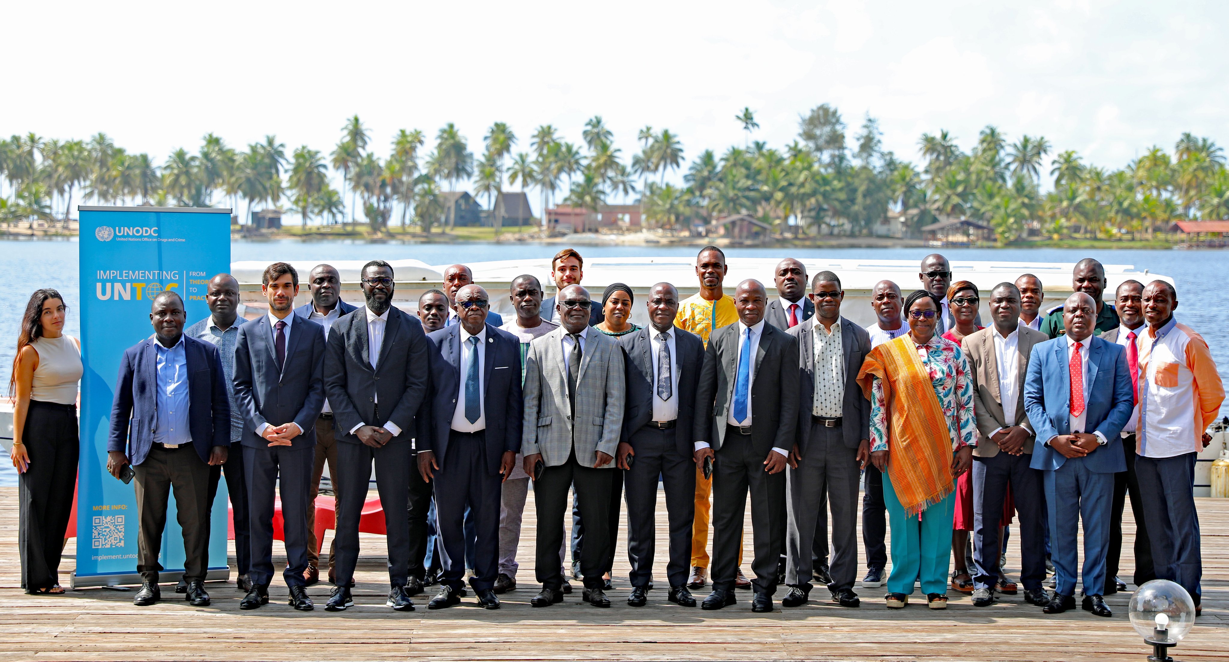A group photo with participants of a meeting taken outside. On the left side of the photo, the roll up of the Global Programme on Implementing the Organized Crime Convention is visible. In the background, palm trees, several houses and the sea.