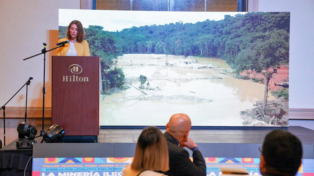 A woman is speaking behind a lectern. Behind her, a screen displays an image showing land covered with water and trees in the back, illustrating the negative effects of illegal mining. Participants of the meeting are sitting on chairs in front of the woman.